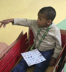 A student sitting in a wagon. A universal core board is attached to a lanyard around his neck.