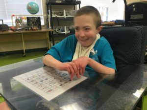 A student sitting in a chair with the 36 location universal core board taped to a tray in front of him.