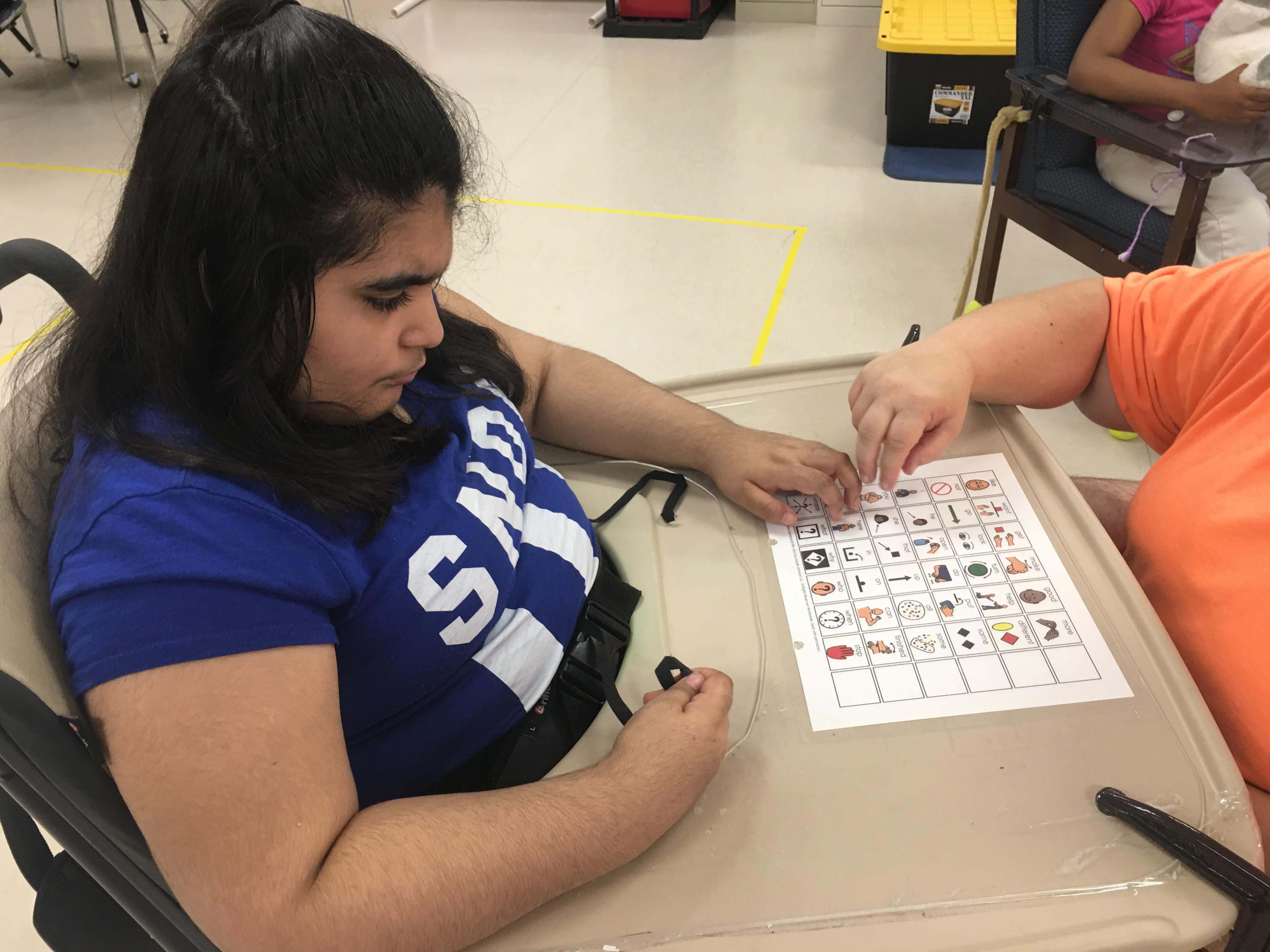 A student pointing at the 36 location universal core board that is taped to the tray of her wheelchair.