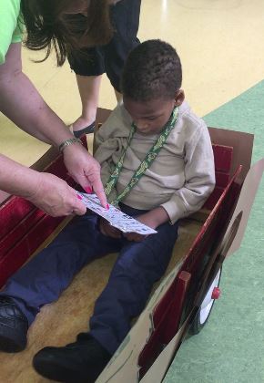 A student sitting in a wagon. An adult is holding a universal core communication book in front of him and pointing at it.